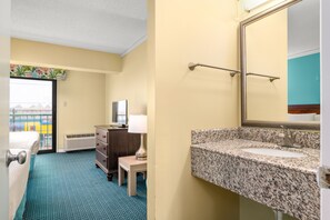 A hotel room with a bed, dresser, lamp, and a bathroom with a granite countertop and mirror. - Cayman Suites Hotel (Ocean City)
