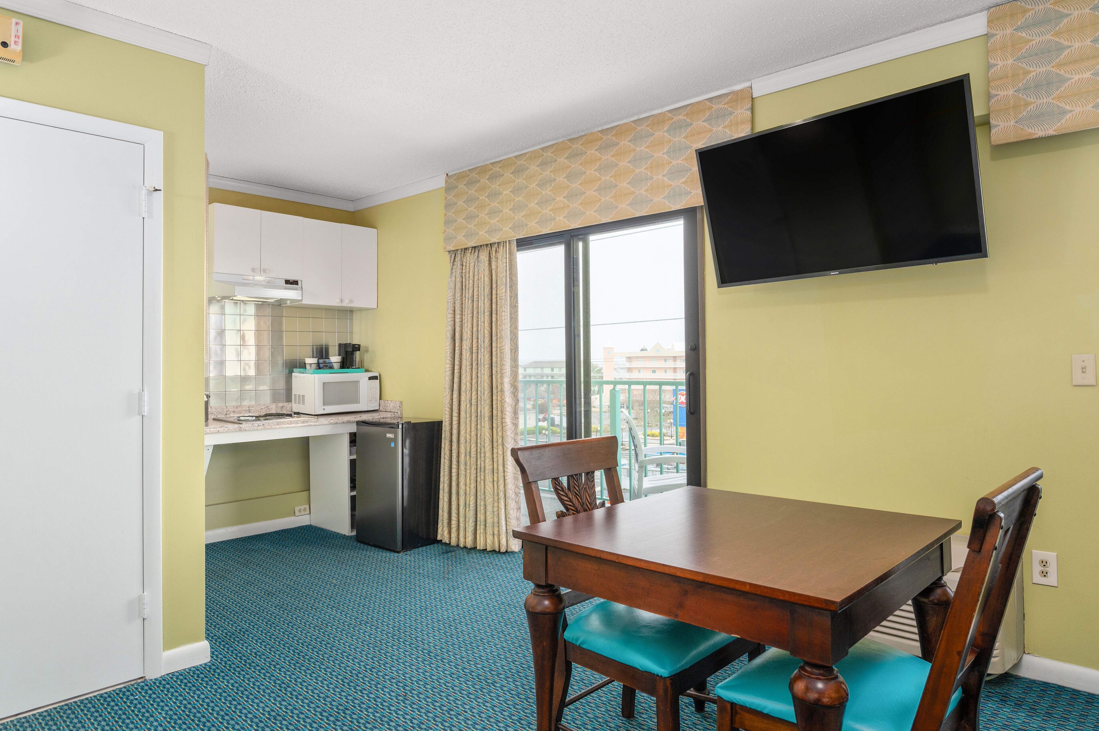 A compact hotel room with a kitchenette, a dining table with chairs, a TV mounted on the wall, and a window with curtains.