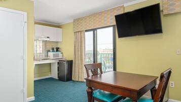 A compact hotel room with a kitchenette, a dining table with chairs, a TV mounted on the wall, and a window with curtains.