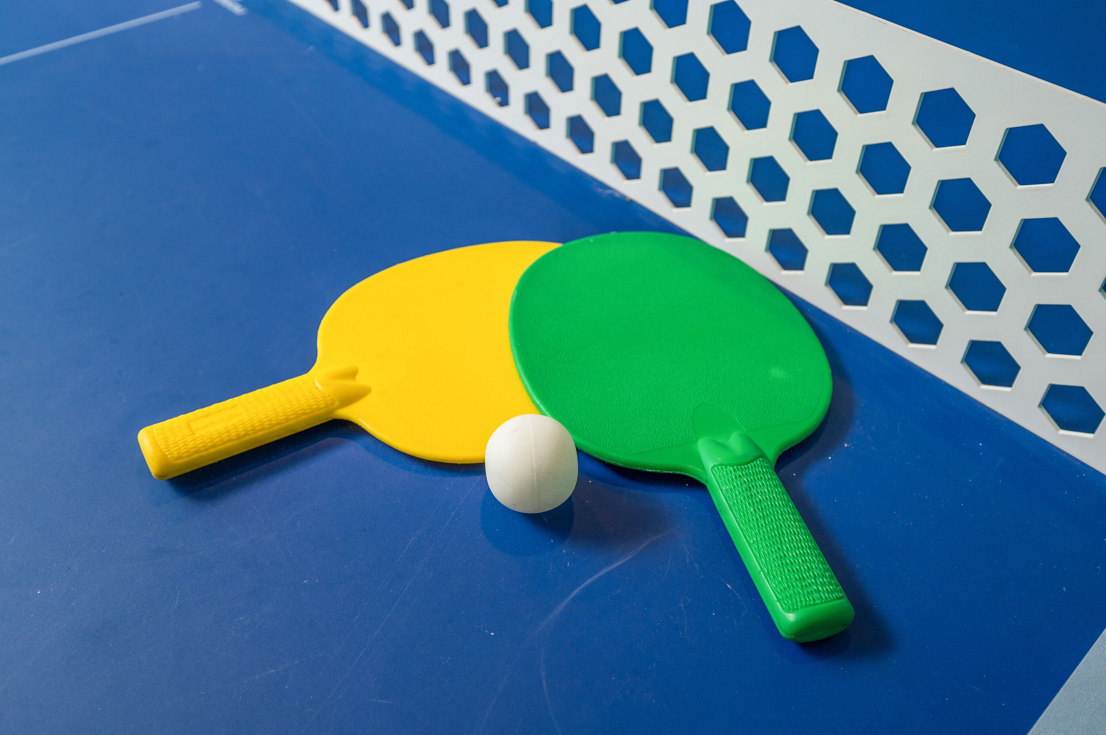 A blue ping pong table with two paddles, one yellow and one green, and a white ball.