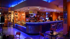 A bar area with a curved counter, bar stools, and a bartender. There are tables and chairs for seating, and the space is well-lit with ceiling lights. - Hotel Blue Rose's (Mesagne)