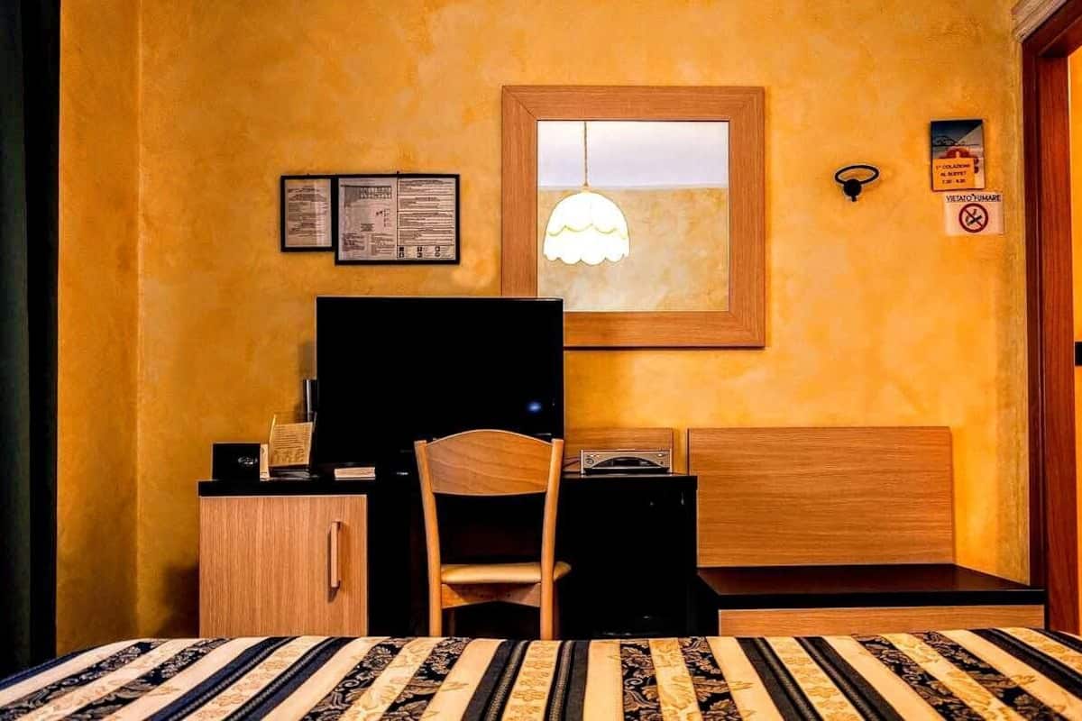 A bedroom with a bed, a desk with a computer, a chair, and a window with a hanging light fixture.