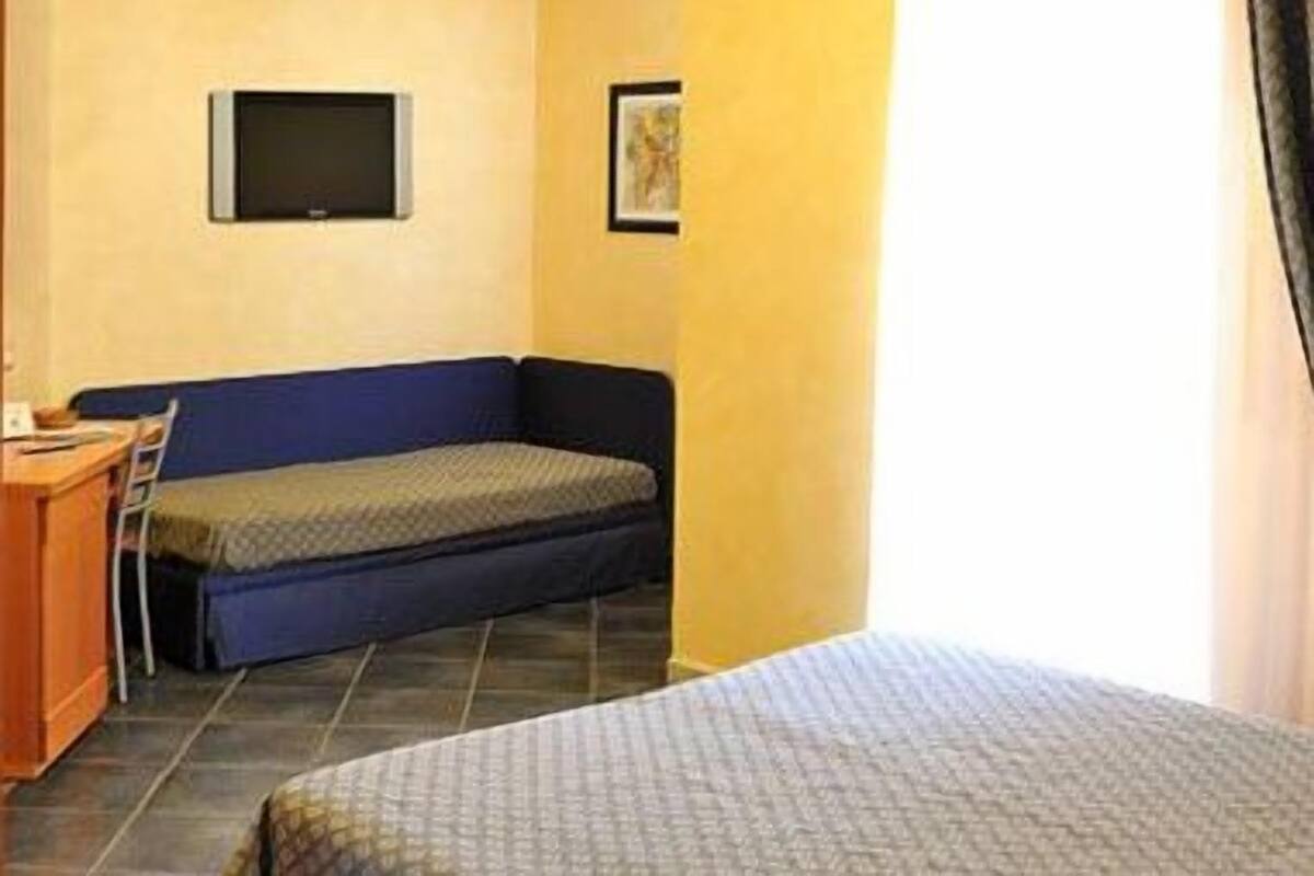 A small room with a bed, a desk, and a chair. There is a television on the wall and a window with curtains.