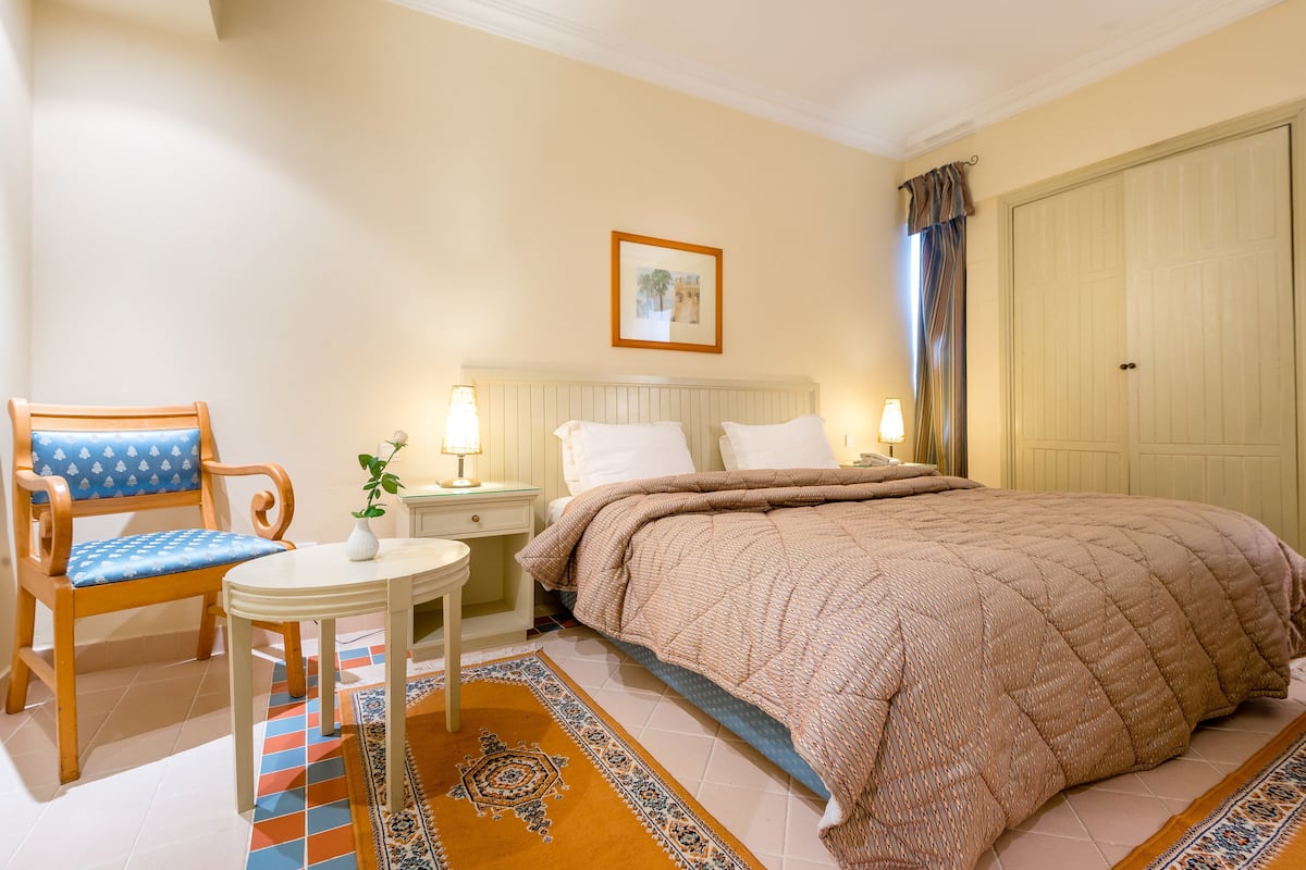 Une chambre d’hôtel avec un lit, une chaise, une table de chevet, une lampe, un tableau au mur et une porte.