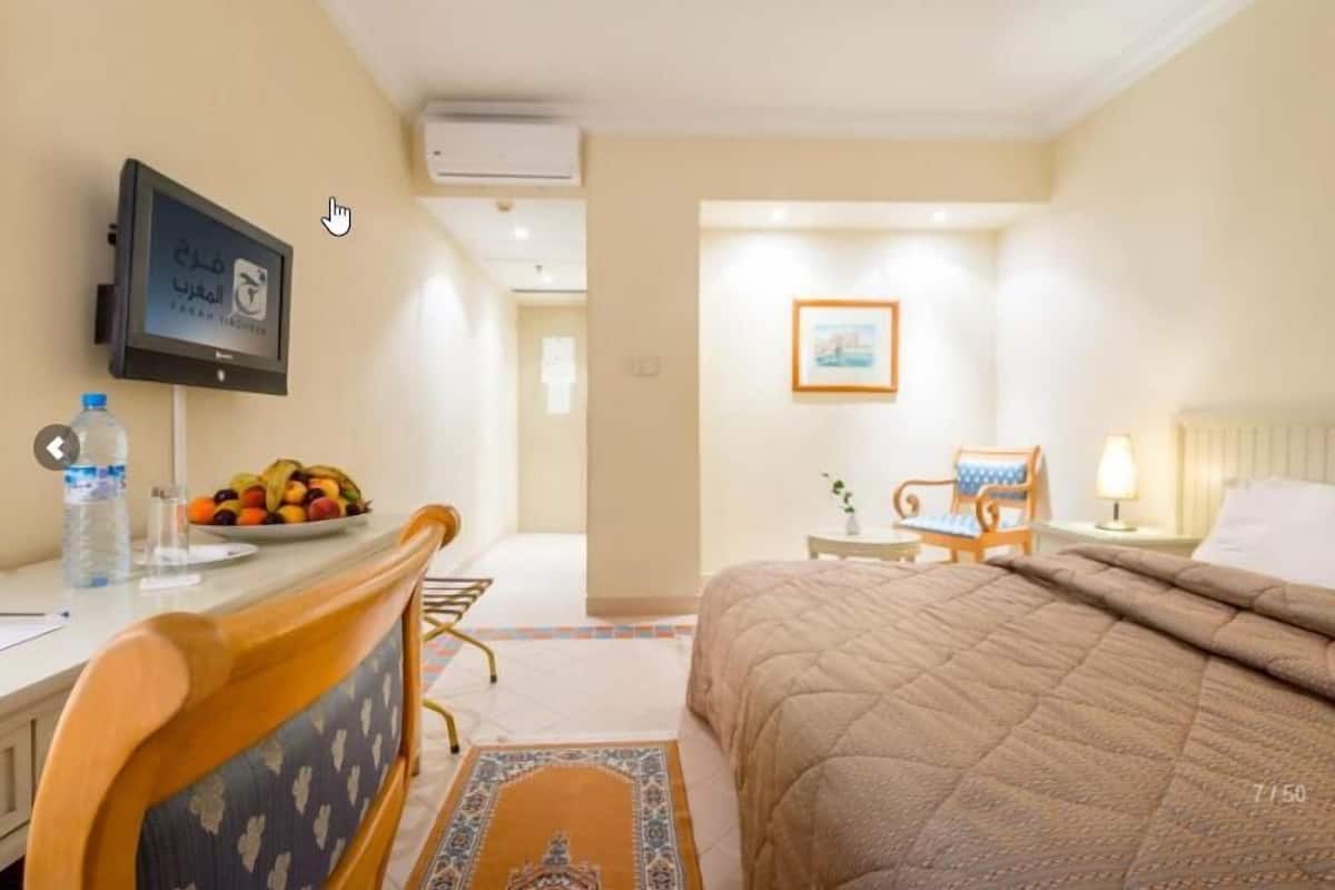 Une chambre d’hôtel avec un lit, un bureau doté d’un écran d’ordinateur, une chaise, une petite table avec une lampe et un tableau encadré au mur.