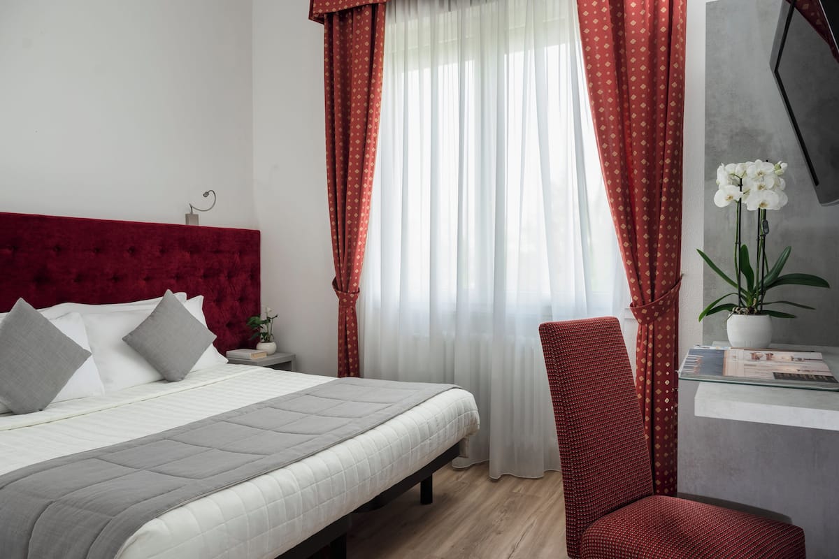 A neatly made bed with a grey quilt, a red upholstered headboard, and a red chair with a patterned cushion. A white orchid plant is on a small table beside the bed.