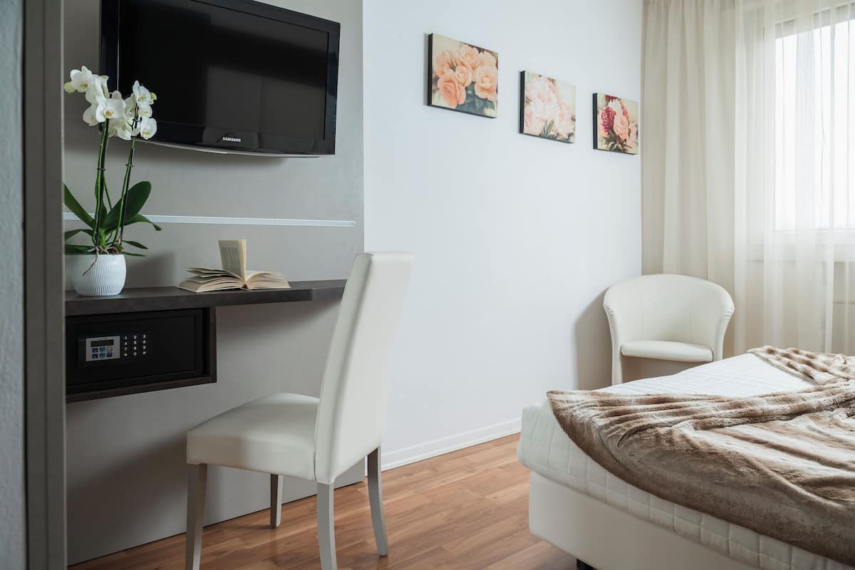 A modern hotel room with a bed, a desk with a chair, a TV mounted on the wall, and a window with curtains.