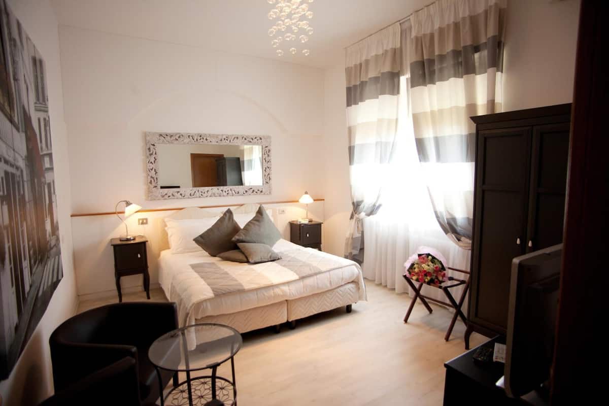A hotel room with a bed, bedside tables, a chair, a small table, a mirror, curtains, and a television.