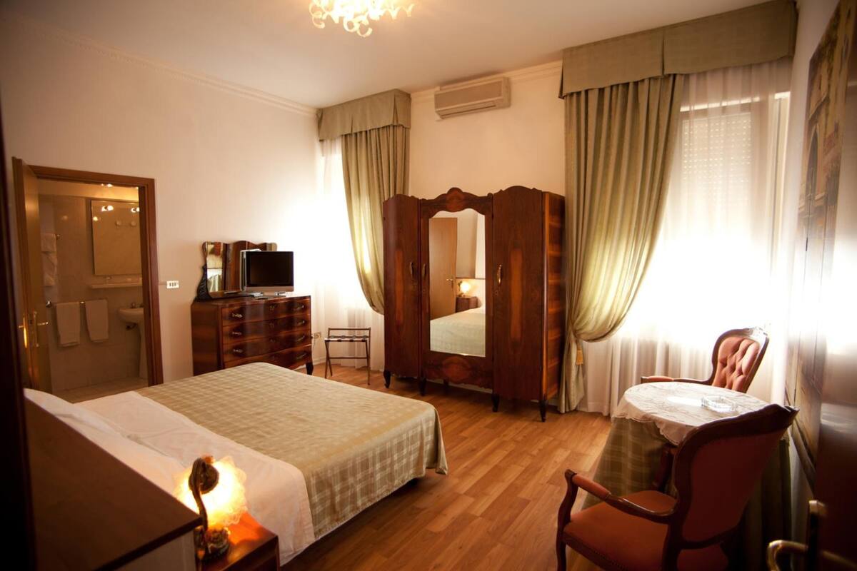 A hotel room with a bed, a desk with a chair, a wardrobe, a television, and a mirror.