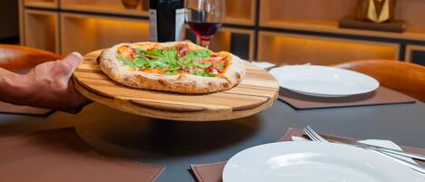 Una pizza sobre una tabla de madera, una botella de vino y una copa de vino tinto sobre una mesa.