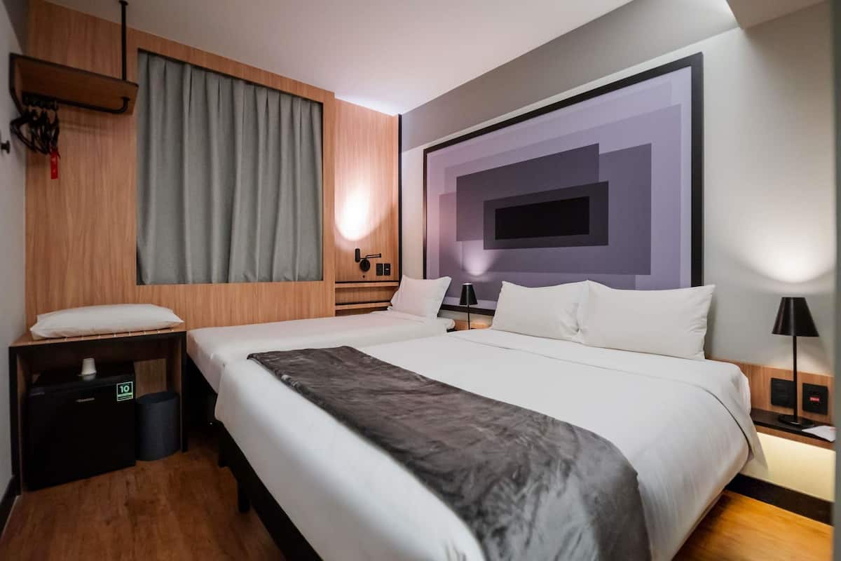 A modern hotel room with a large bed, wooden headboard, grey curtains, a wall-mounted TV, and a bedside table with a lamp.