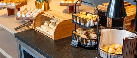 A buffet counter with various baked goods, including pastries and bread, arranged on wooden stands and wire baskets.
