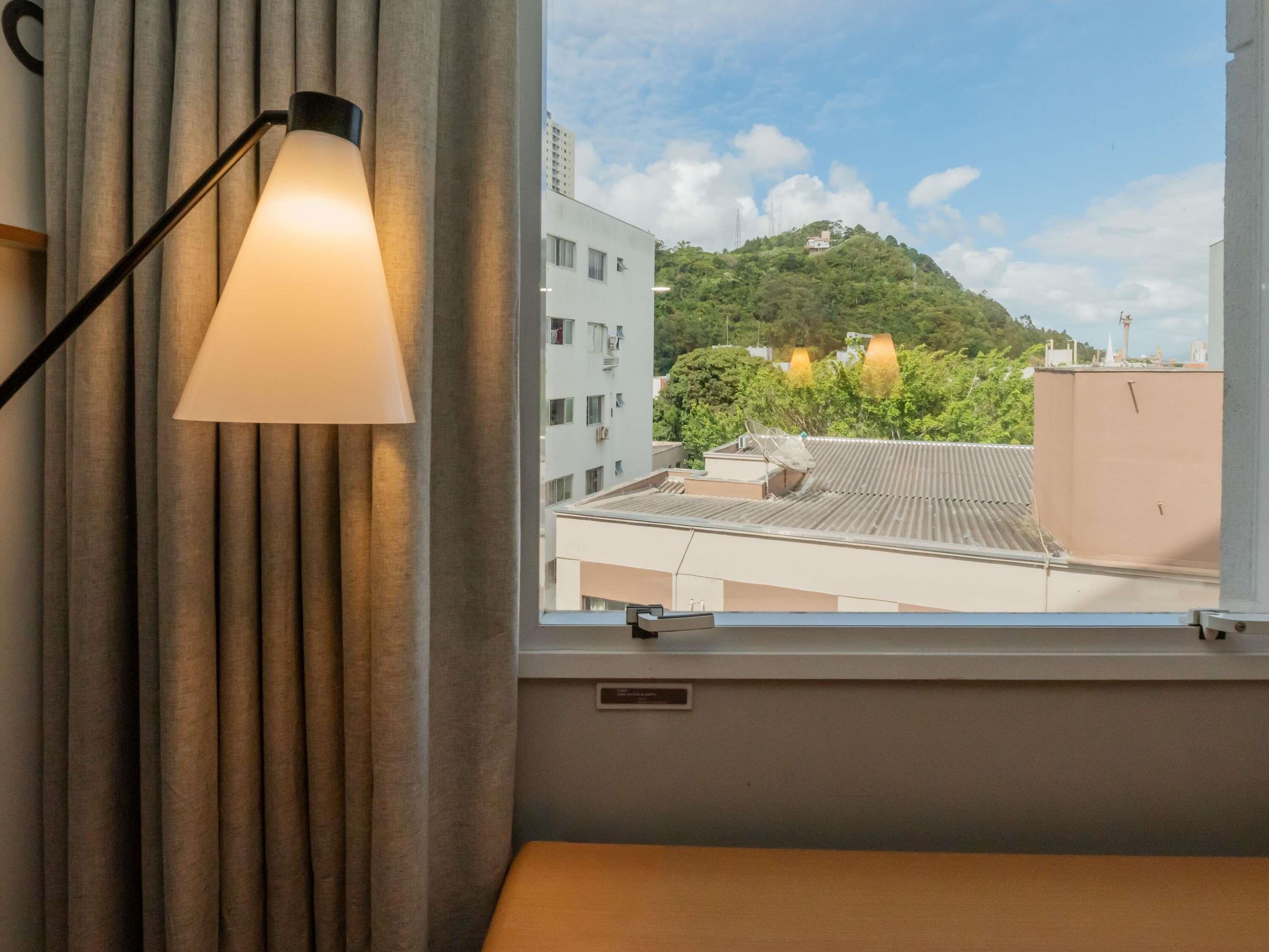 Een kamer met een raam, een lamp en uitzicht op gebouwen en een heuvel.