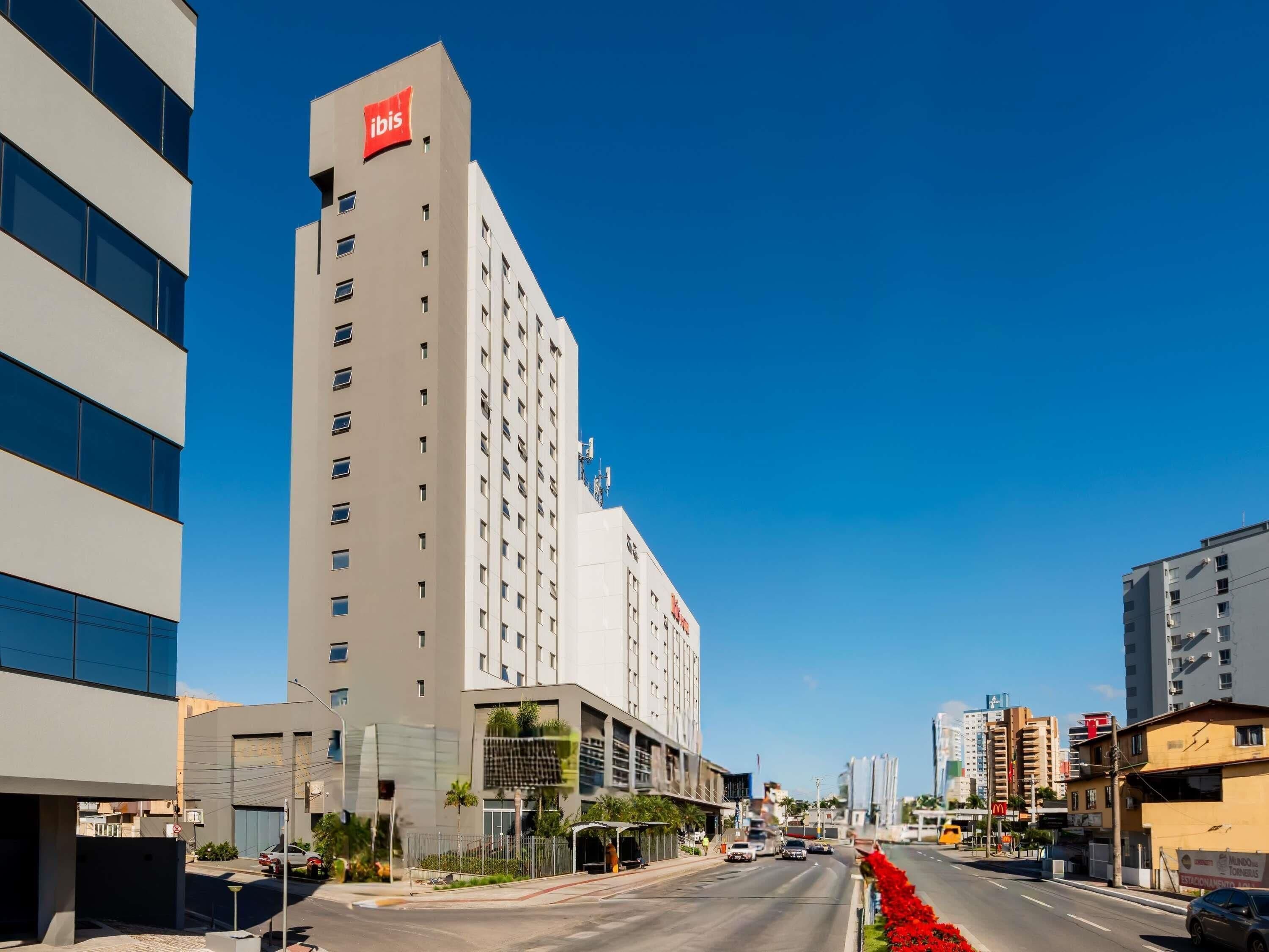 A modern hotel with a prominent red logo, multiple floors, and a clear view of the street in the background.