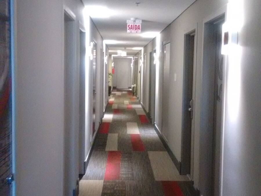 Corredor de hotel com carpete quadriculado, portas em ambos os lados e placa 'SAÍDA' suspensa.