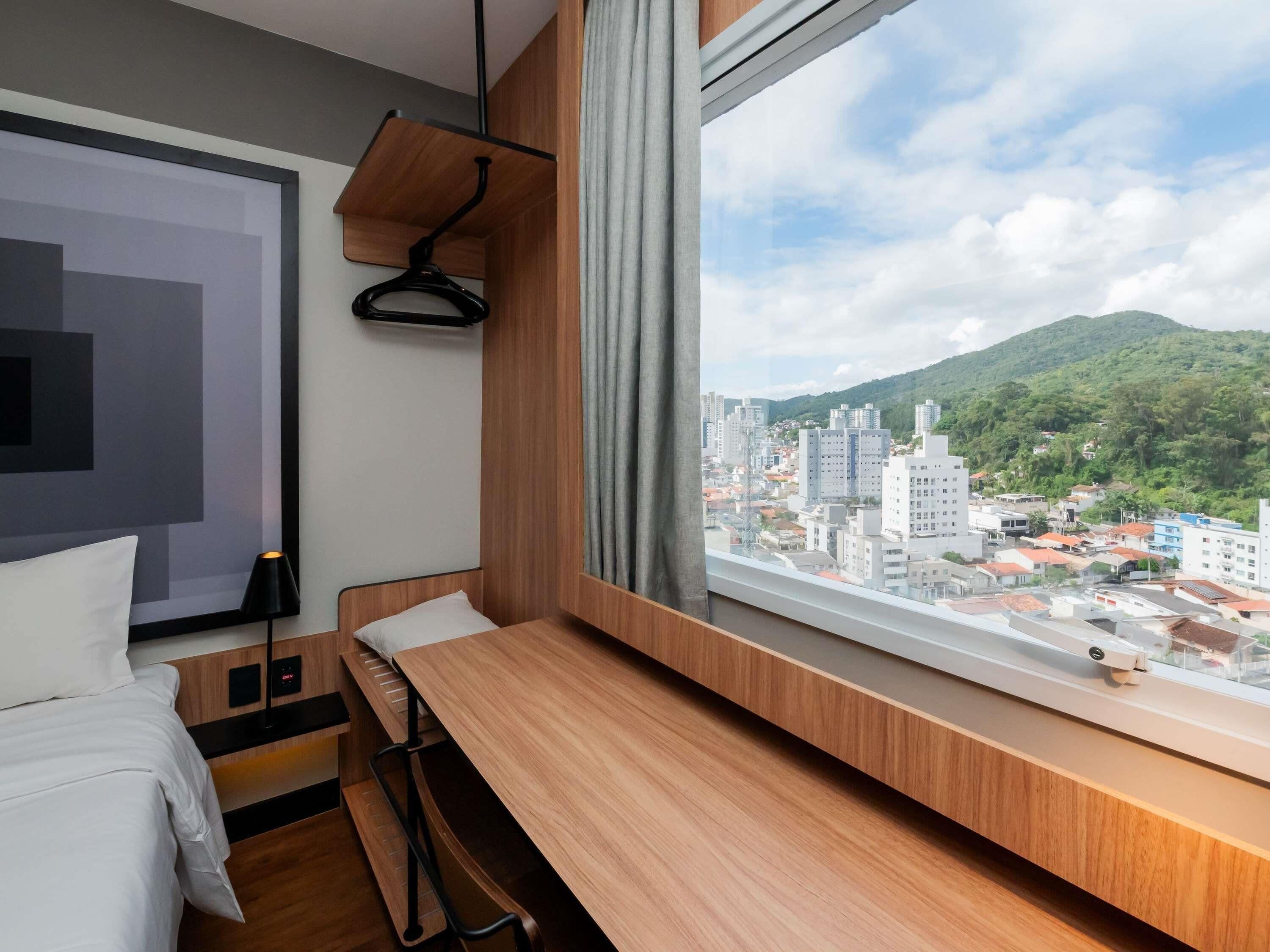 A modern hotel room with a wooden desk, a bed, a hanging light, and a view of a cityscape and mountains.