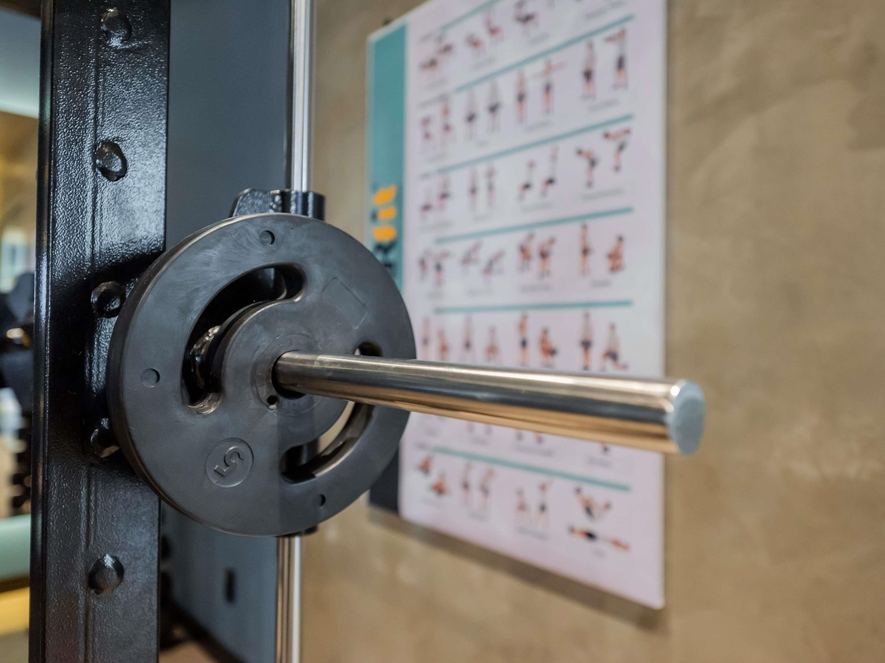 Ein Krafttrainingsgerät mit einer Langhantel, ein an der Wand befestigtes Trainingsposter und ein metallener Griff.