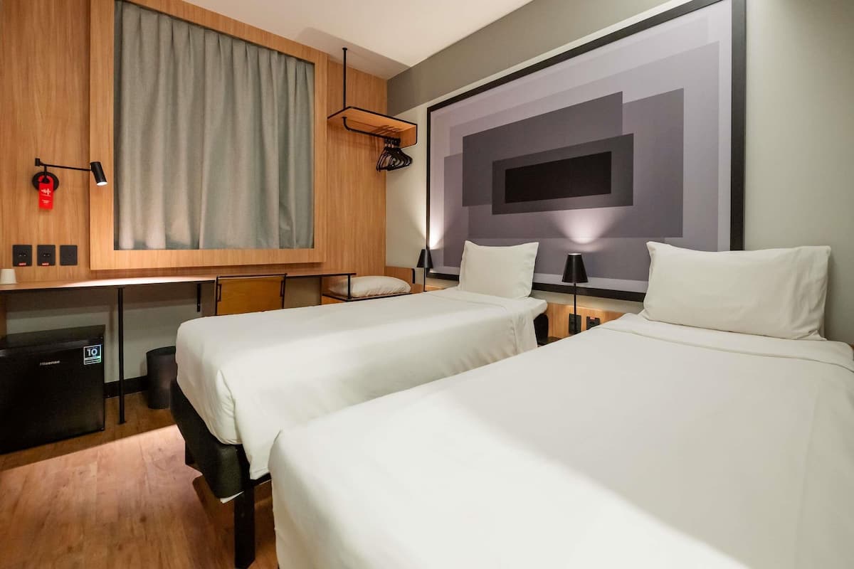 A hotel room with two beds, a desk, a chair, a TV, and a large window with curtains.