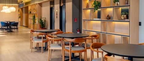 A modern interior with round tables and chairs, wooden shelving with plants, and a visible fire alarm.