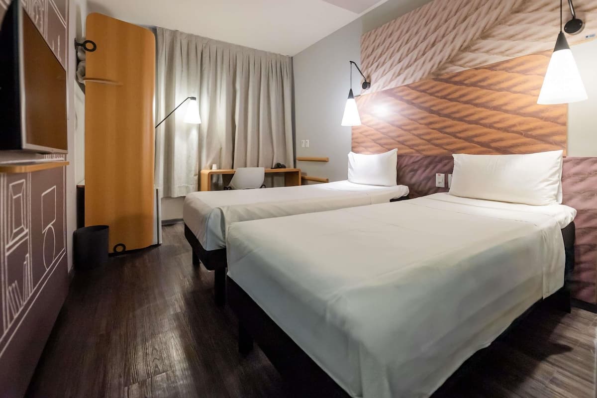 A hotel room with two beds, a desk, a chair, a TV, and a patterned wall.