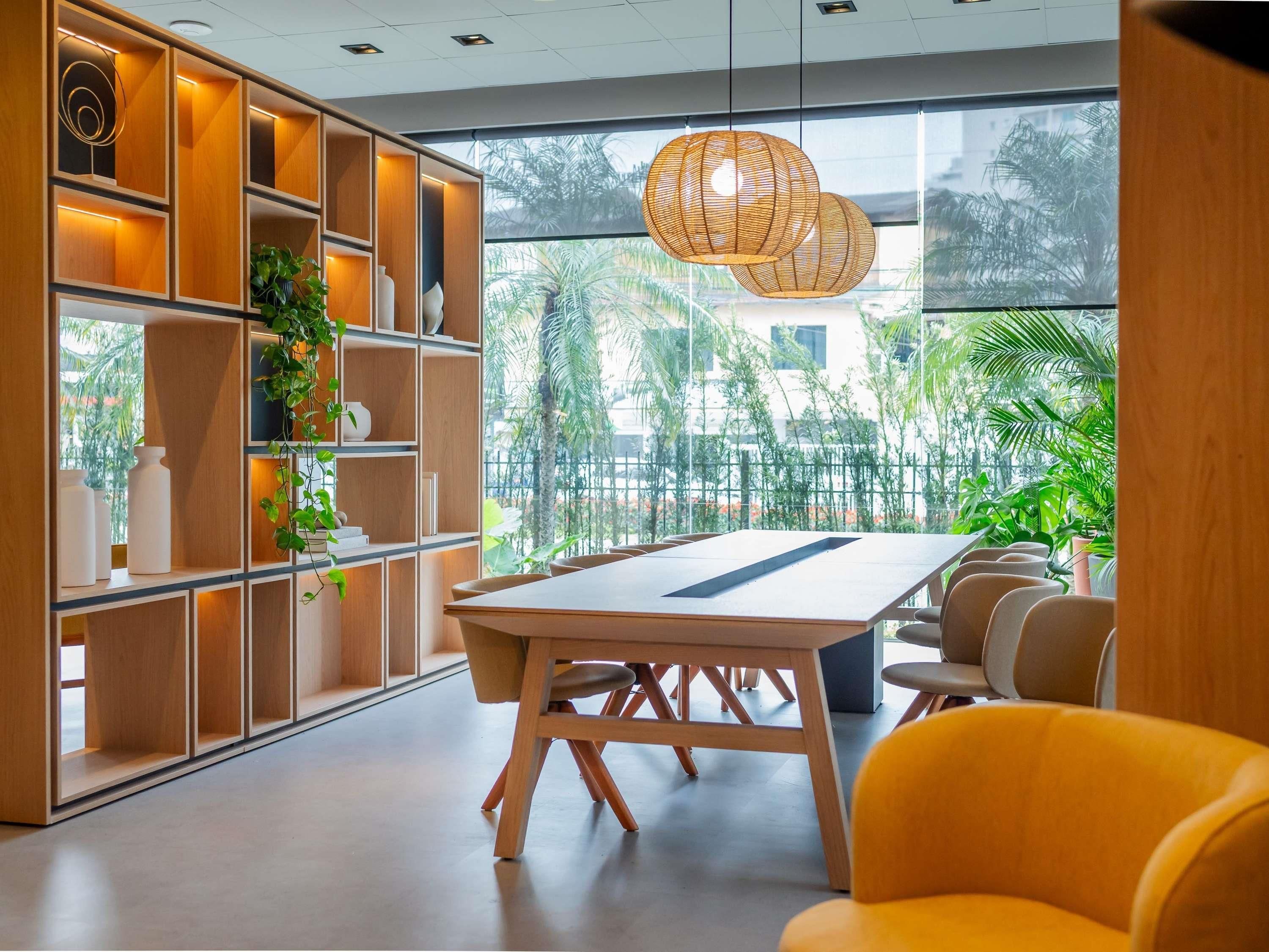 Un intérieur moderne avec une grande table et des chaises, des étagères en bois ornées de plantes, et une vue sur la verdure à l’extérieur.
