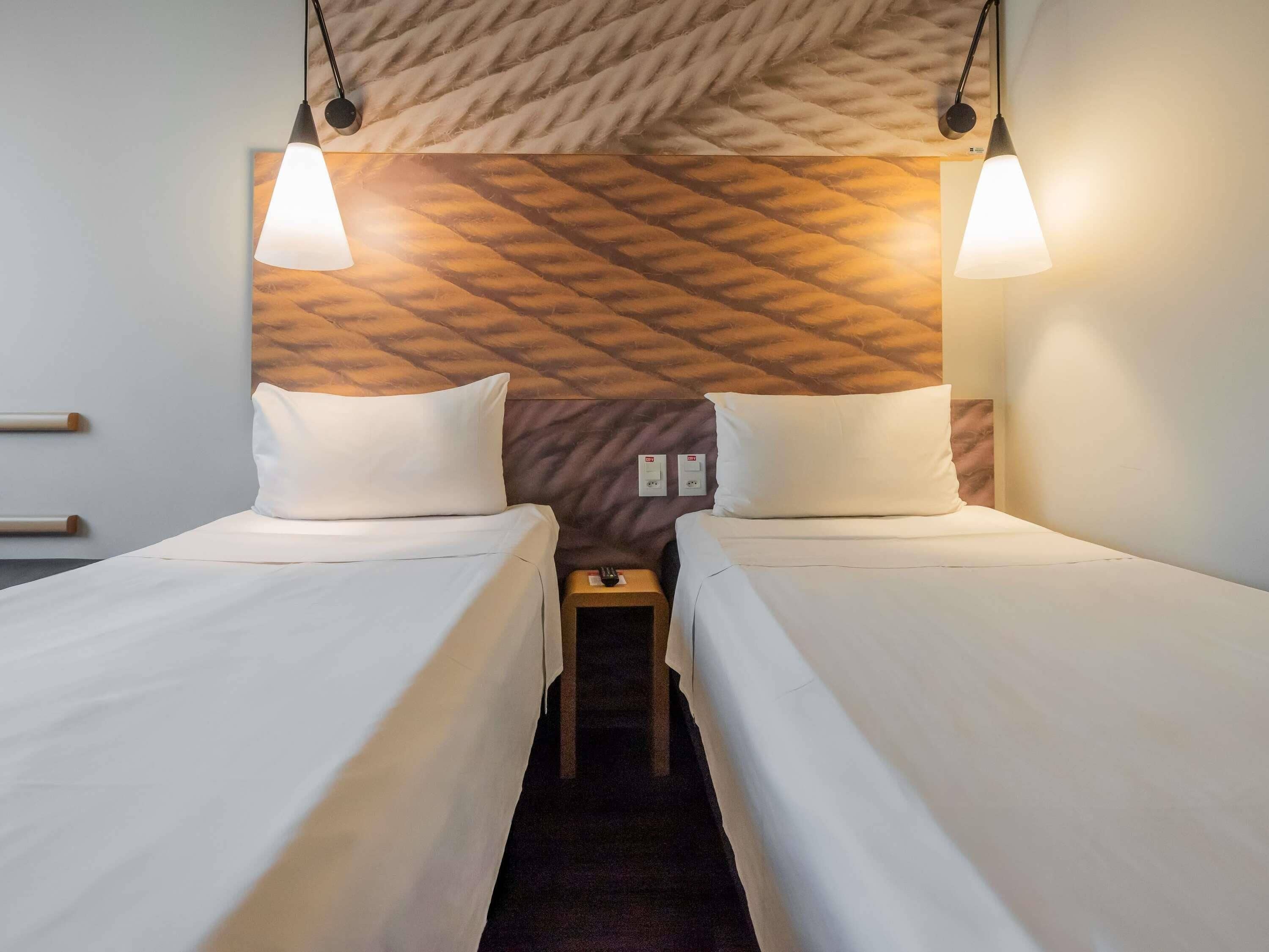 A hotel room with two beds, a headboard with a geometric pattern, and pendant lighting.