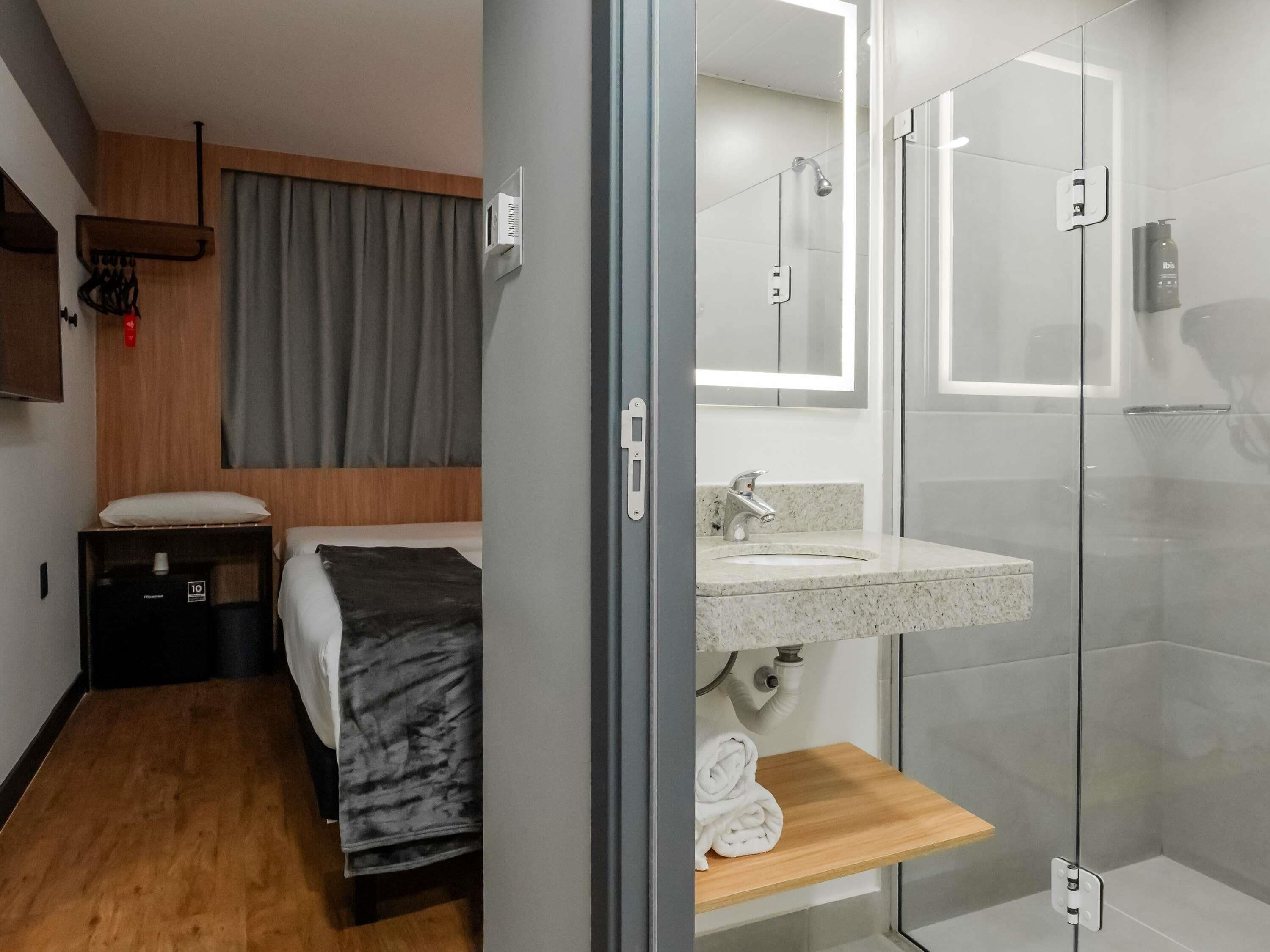 Una habitación de hotel moderna con una cama, un pequeño estante de madera, un baño con cabina de ducha de vidrio y un lavamanos.