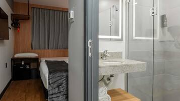 Una habitación de hotel moderna con una cama, un pequeño estante de madera, un baño con cabina de ducha de vidrio y un lavamanos.