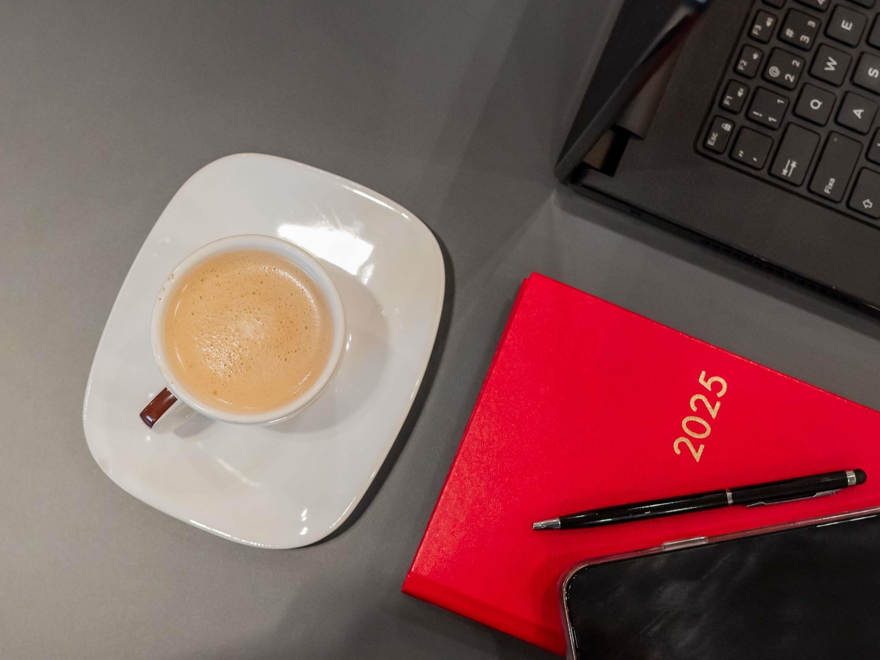Eine Kaffeetasse auf Untertasse, ein rotes Notizbuch mit der Aufschrift '2025', ein Stift und ein Smartphone auf einem Tisch.
