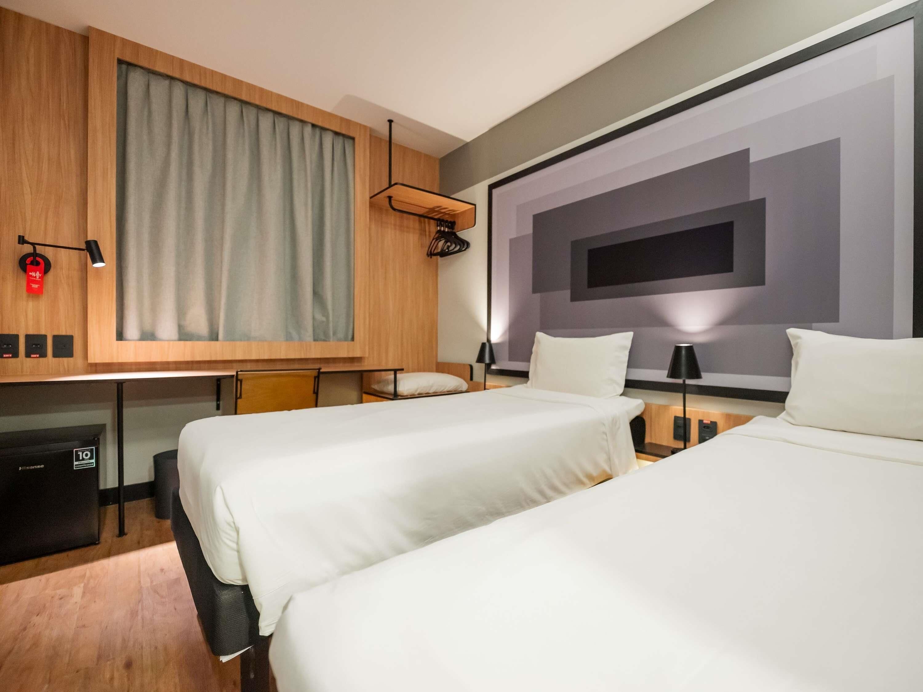 A hotel room with two beds, a desk, a minibar, a large window with curtains, and a wall-mounted TV.