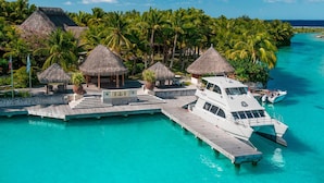 Un resort tropical con cabañas de techo de paja, un yate blanco amarrado en un muelle y un mar turquesa cristalino.