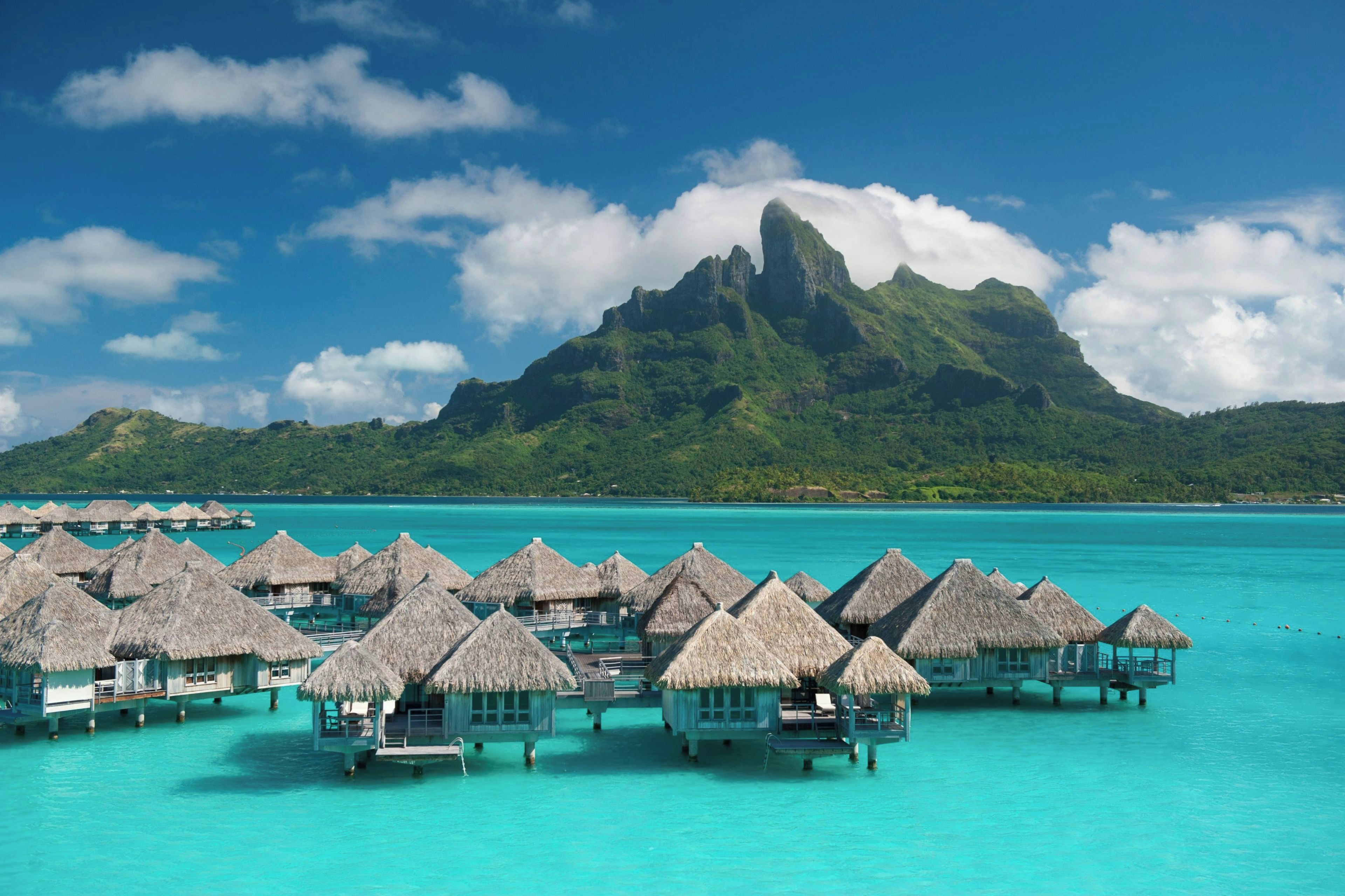 Wasserbungalows mit Strohdächern, ein klares türkisfarbenes Meer und ein markanter Berg im Hintergrund.