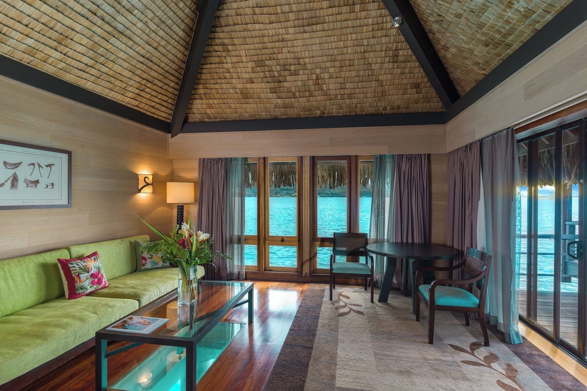Um quarto com teto de madeira, um sofá verde, uma mesa de centro de vidro, uma mesa de jantar com cadeiras e grandes janelas com cortinas.