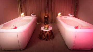 A spa room with two white tubs, a small wooden table with candles, and a curtain backdrop.