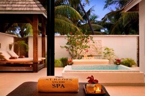 A spa area with a massage table, a towel with 'Stregis Bora Bora Spa' text, a tray with a drink and candle, and a pool with a view of palm trees.