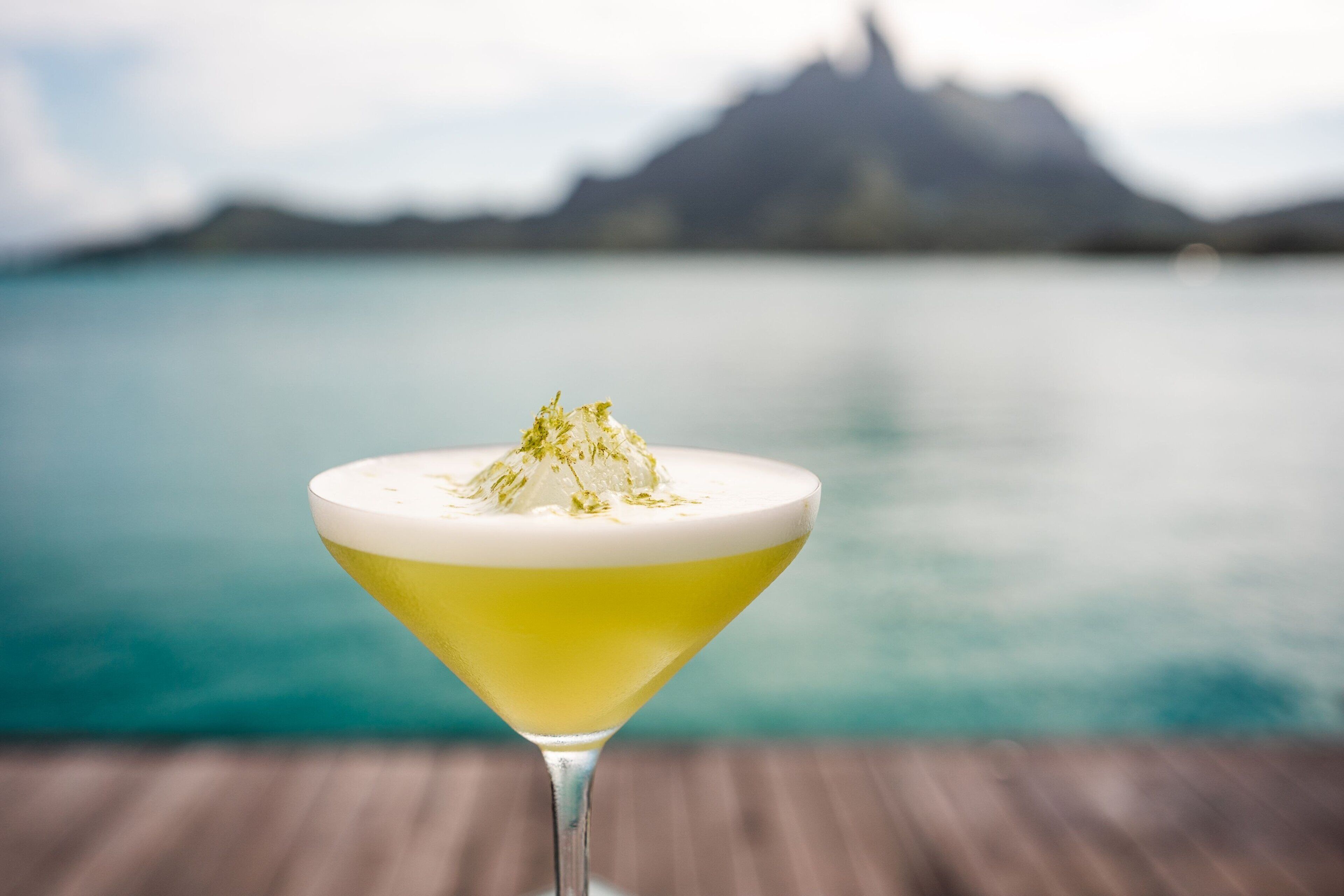 Coquetel num copo de martini com cobertura cremosa, com um corpo d'água calmo e montanhas ao fundo.