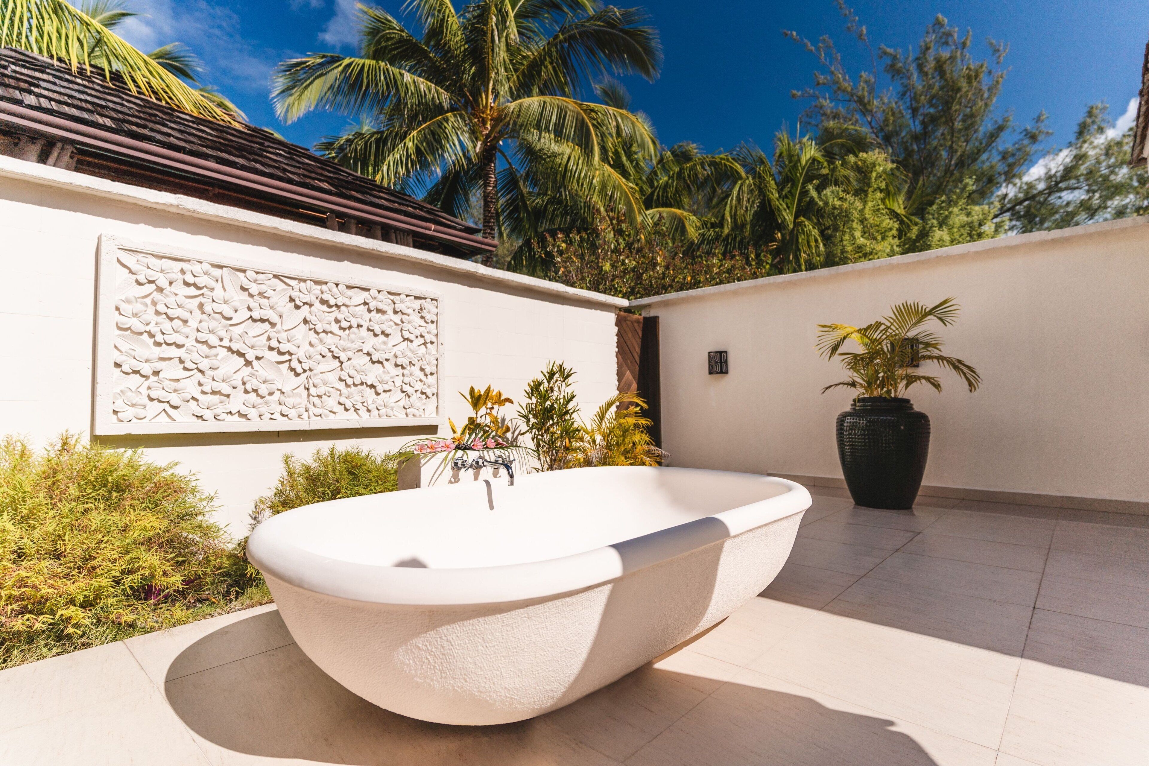 A modern outdoor bathroom with a freestanding tub, surrounded by lush greenery and palm trees.