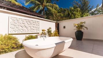A modern outdoor bathroom with a freestanding tub, surrounded by lush greenery and palm trees.