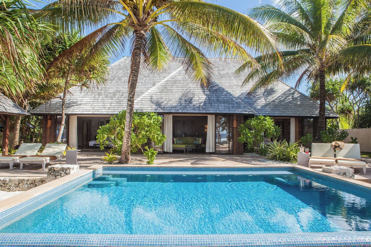 Área de piscina com espreguiçadeiras e um espaço coberto para relaxar, cercada por palmeiras e um edifício com telhado de azulejos.