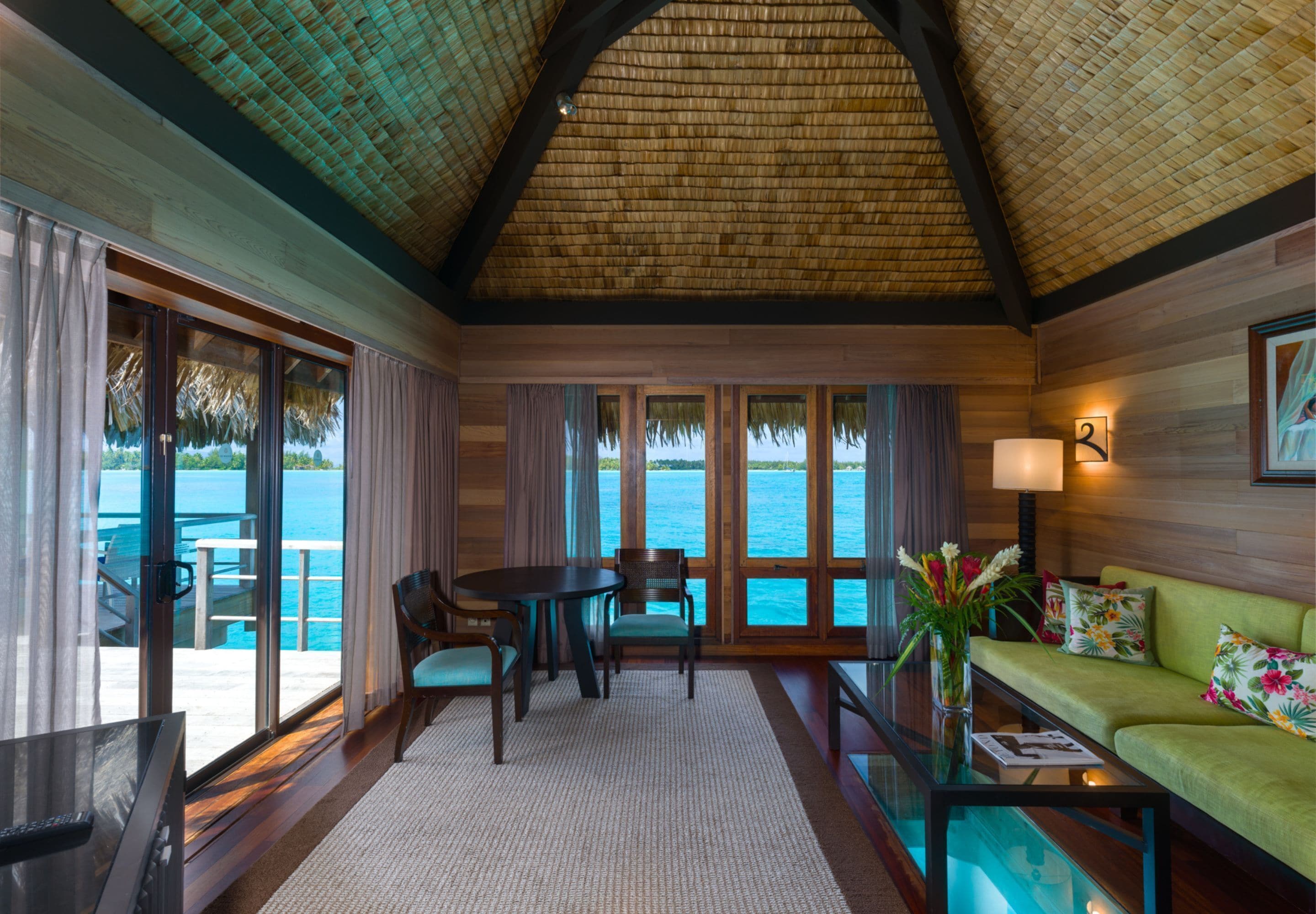 A room with a large window overlooking a body of water, a dining table with chairs, a sofa, and a TV.