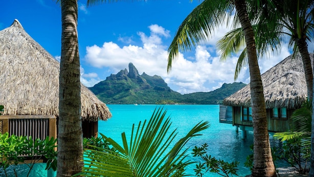 Un resort tropical con bungalows sobre el agua con techo de paja, un mar turquesa cristalino y una montaña al fondo.