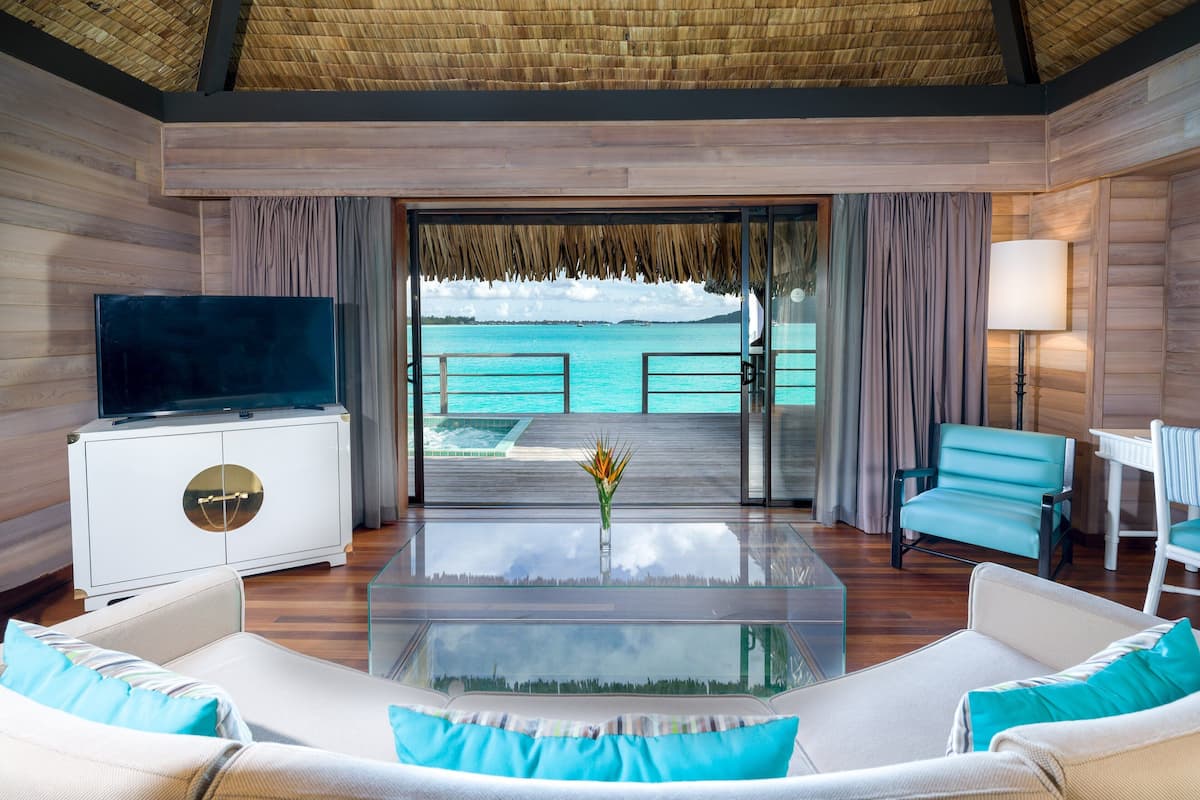 Quarto moderno com mesa de centro de vidro, sofá branco e poltronas azul-petróleo. Uma televisão está fixada em um móvel branco, e há vista para o oceano através de uma porta de vidro.