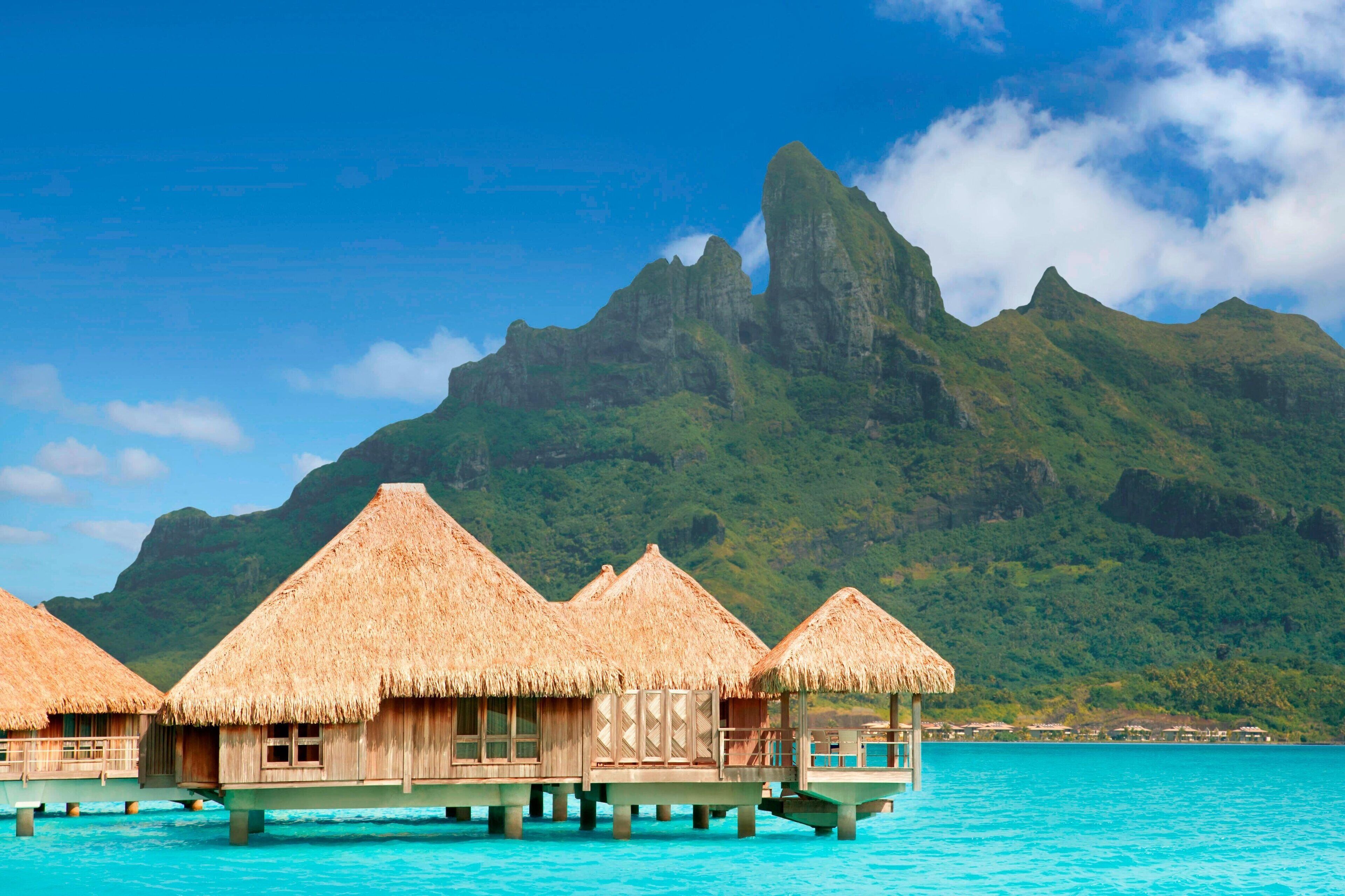 Overwater bungalows with thatched roofs, wooden structures, and a clear view of turquoise waters and a mountain in the background.
