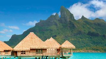Overwater bungalows with thatched roofs, wooden structures, and a clear view of turquoise waters and a mountain in the background.