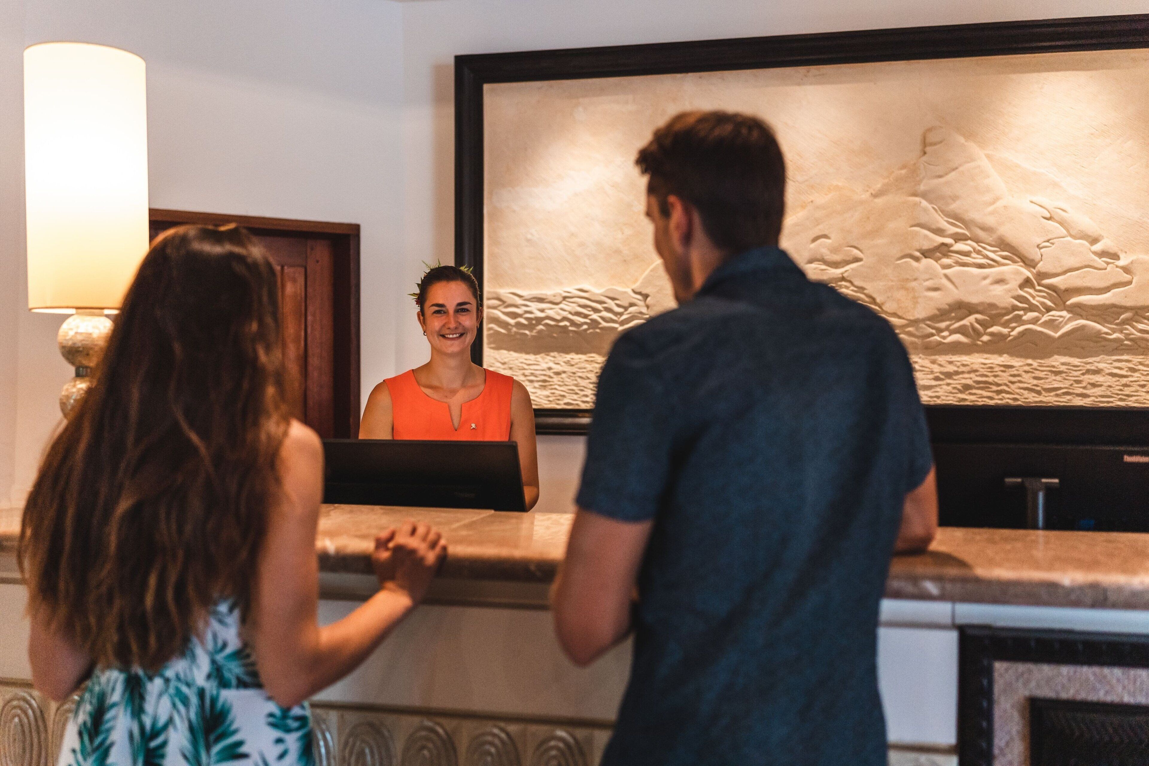 A reception area with a receptionist behind a counter, a large framed artwork on the wall, and a lamp to the left.