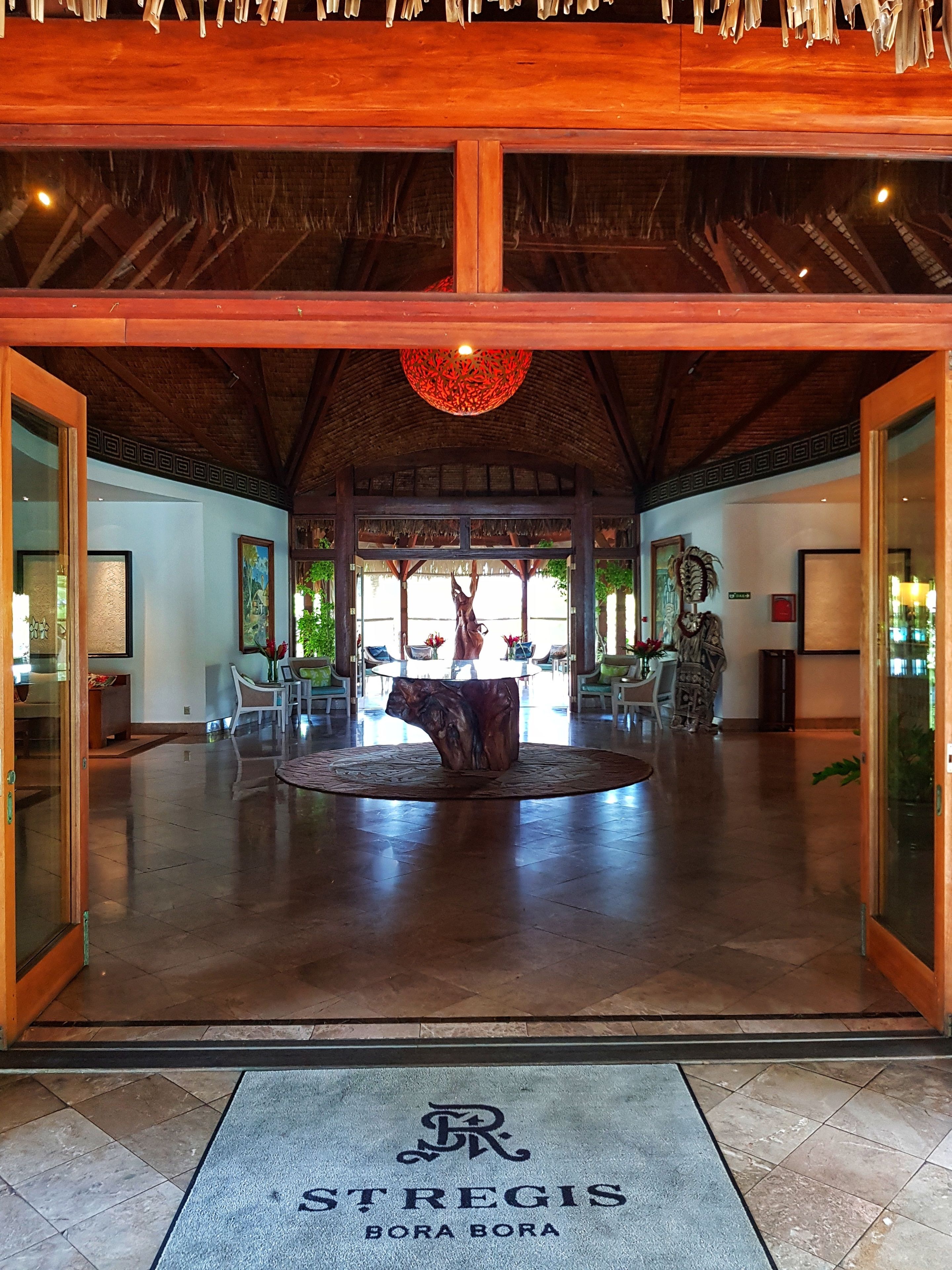 A spacious lobby with a central wooden table, a decorative ceiling light, and a large window offering a view of the outdoors.
