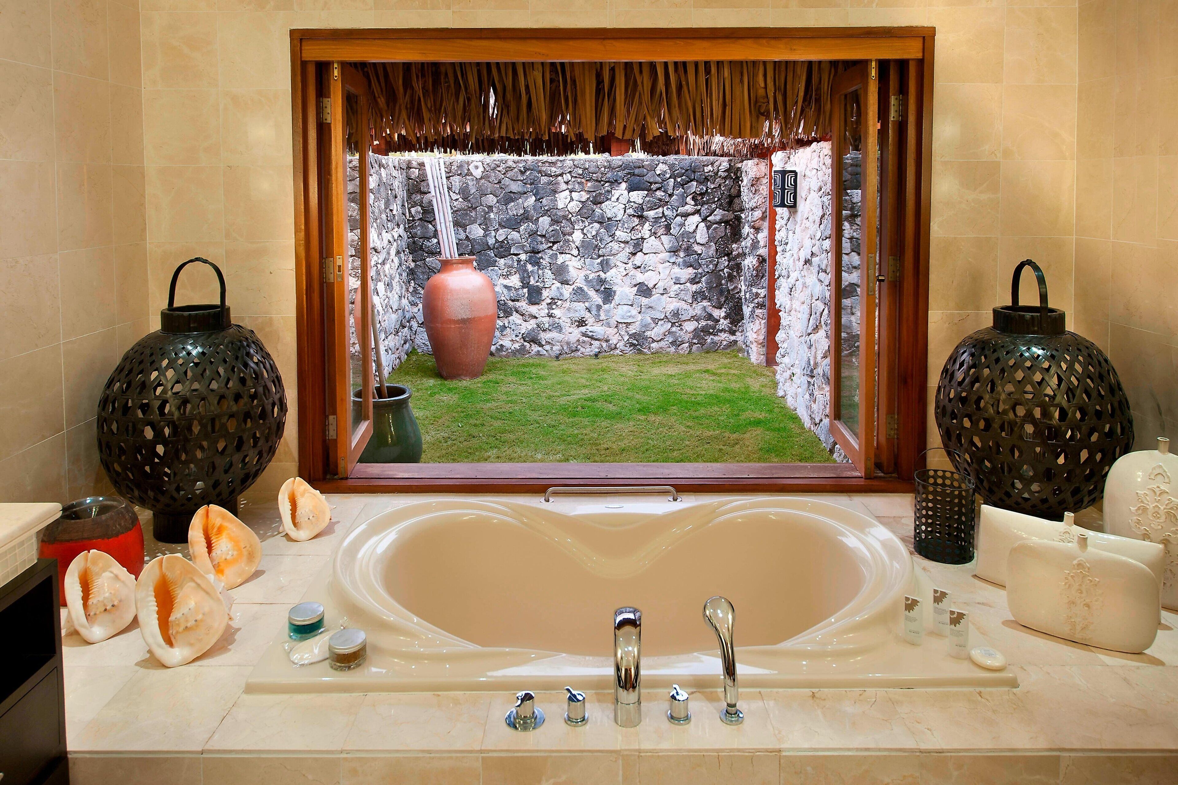 Un baño con una bañera grande, una pared de piedra visible a través de una ventana, y objetos decorativos como jarrones y velas.