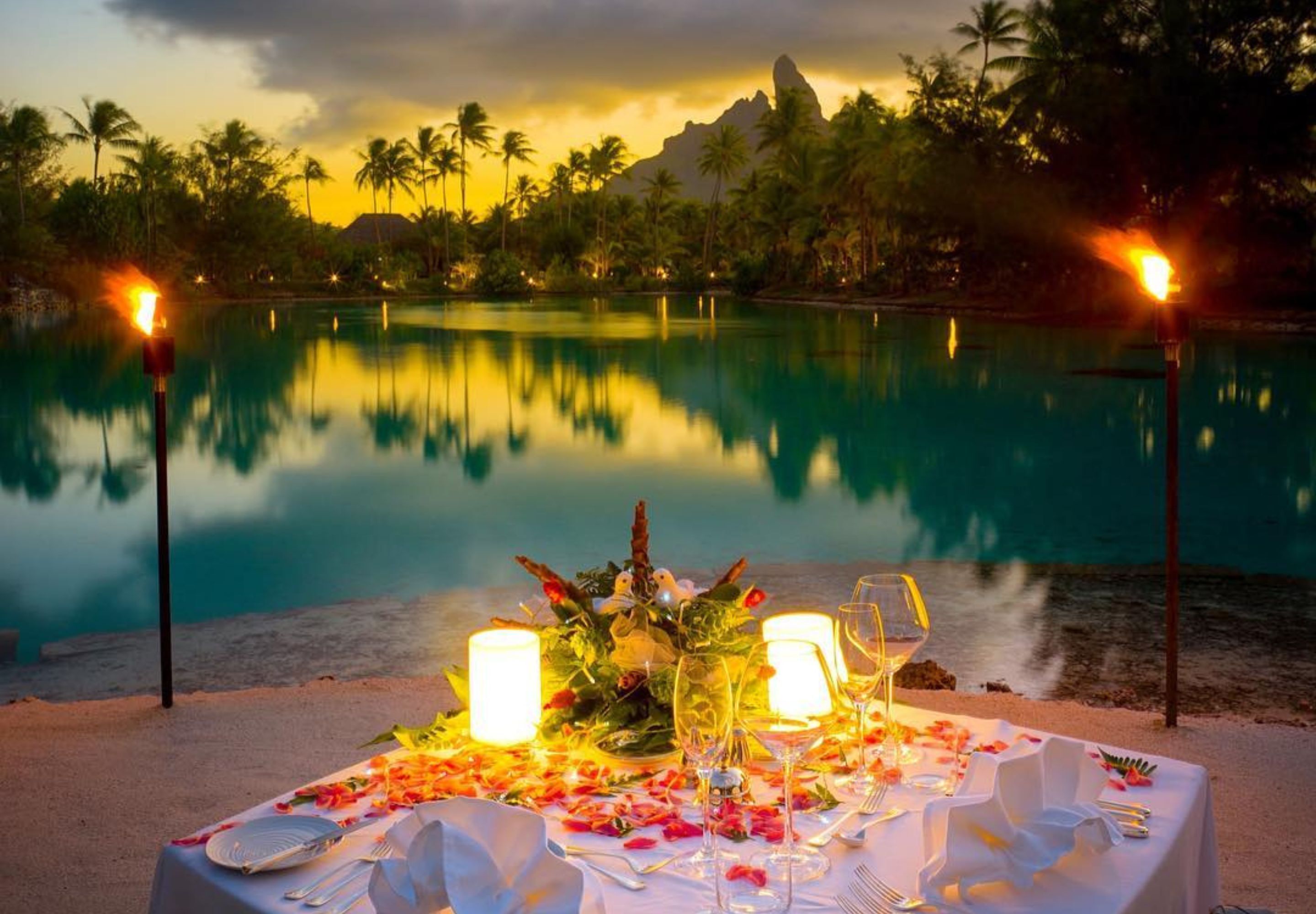 A table set for a meal with candles and glasses, overlooking a calm pool and palm trees.