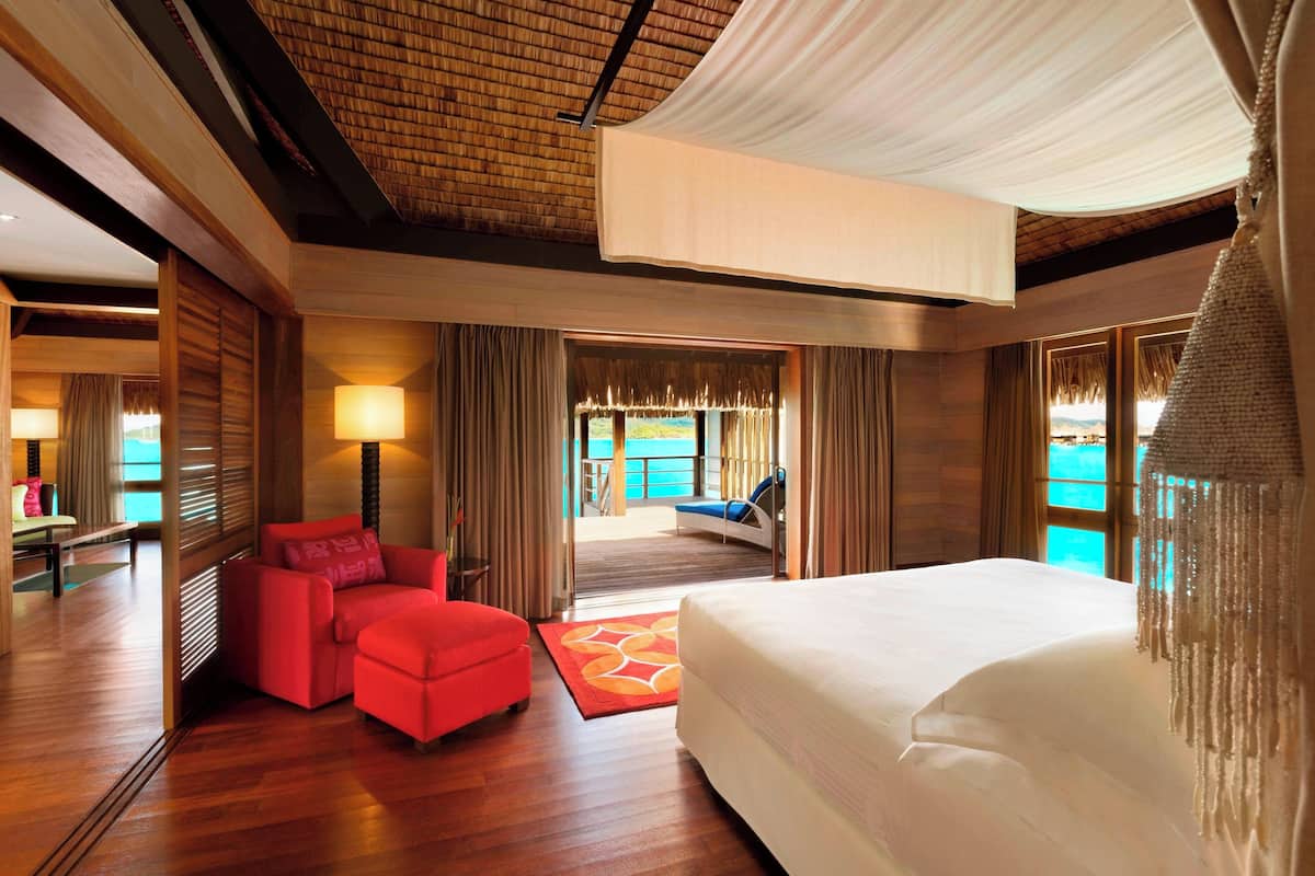 Quarto de hotel com uma cama grande, um sofá vermelho, piso de madeira e vista para o mar pela janela.