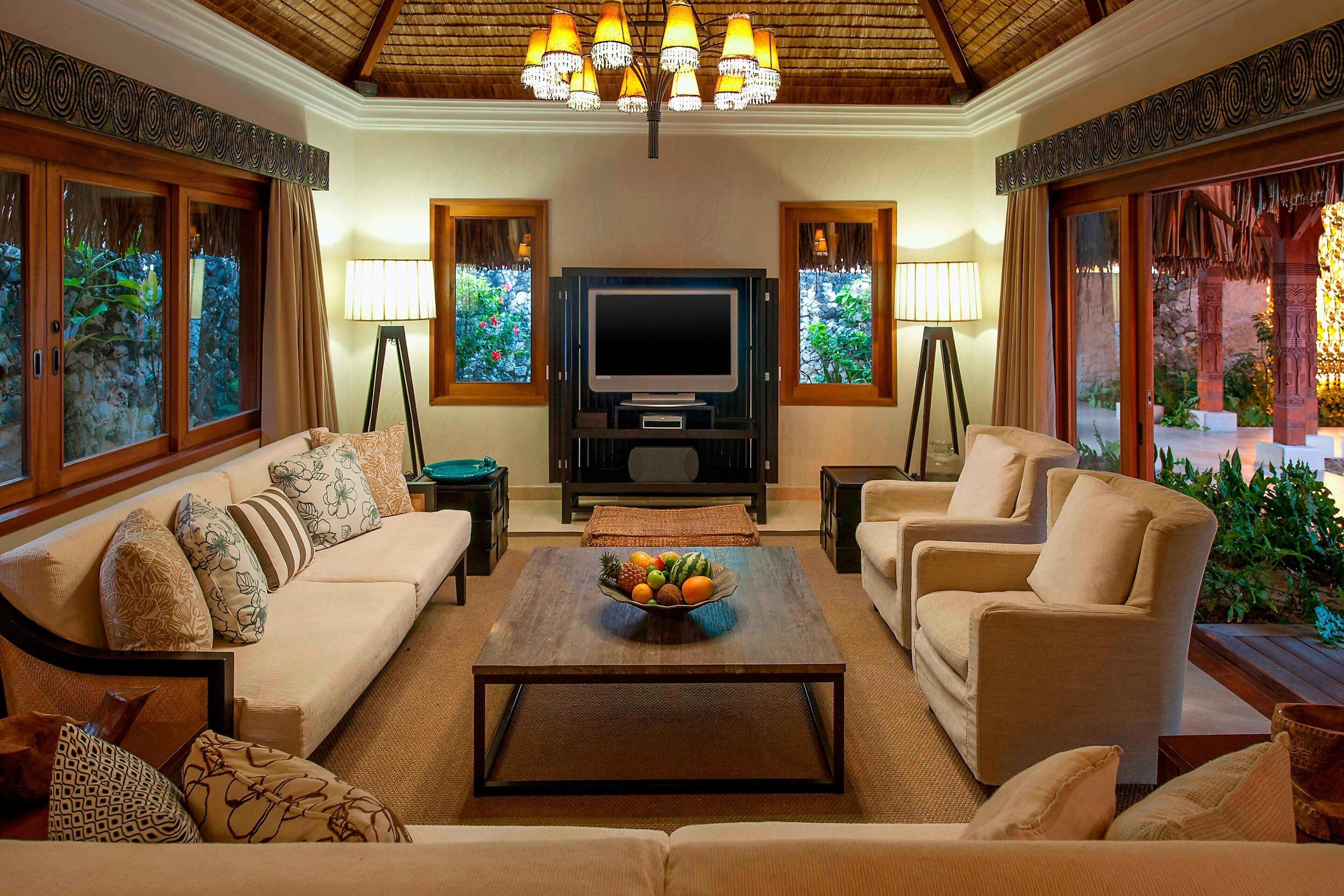 A living room with a sofa, armchairs, a coffee table with fruit, a TV, and decorative lamps.